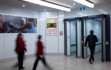 Indoor airport arrivals area with white walls and tiled flooring, featuring glass exit doors marked “Way Out.” A digital advertising screen on the wall displays a “Do more you at Scarborough Beach”